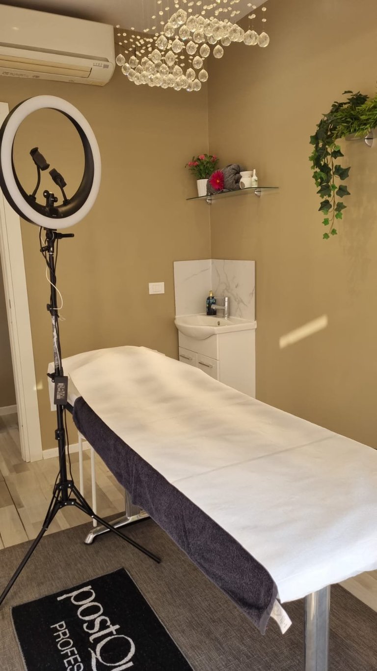 Professional spa treatment room with massage table, ring light, crystal chandelier, potted plants, and beige walls